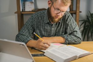 Man studying and taking notes with a Bible on a desk indoors.