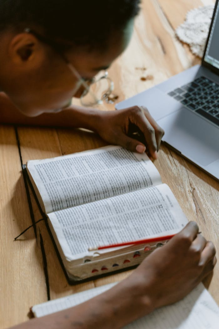 A young adult reading the Bible beside a laptop, deep in study.